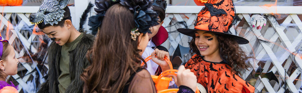 Preteen fille tenant seau près des amis interracial en costumes d'Halloween à l'extérieur, bannière  - Photo, image