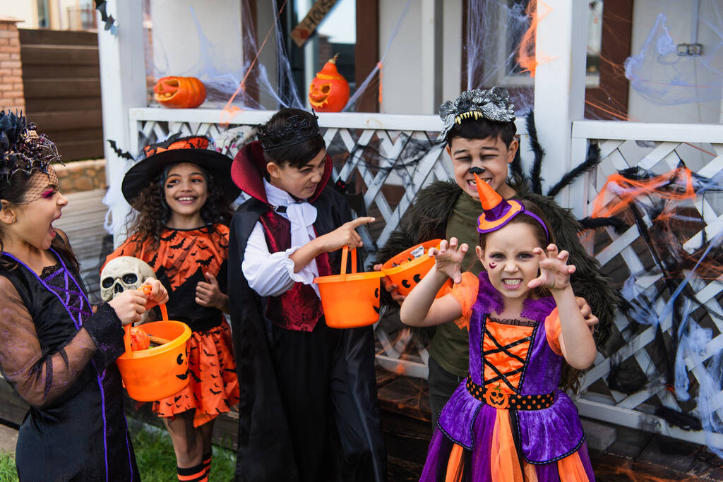 Niños interraciales en disfraces de Halloween sosteniendo cubos y jugando al aire libre  - Foto, imagen