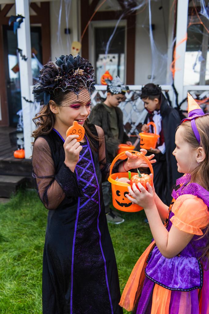 Smiling kid in halloween costume holding candy and bucket near friend in backyard  - Photo, Image