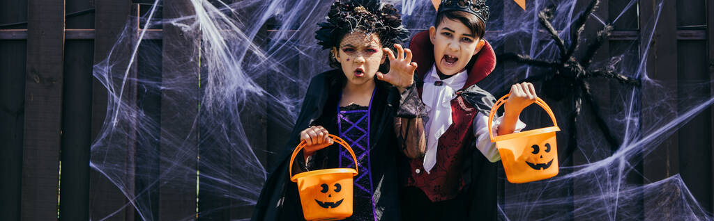 Bambini multietnici che tengono secchi di Halloween e smorfie davanti alla telecamera vicino all'arredamento su recinzione, striscione  - Foto, immagini