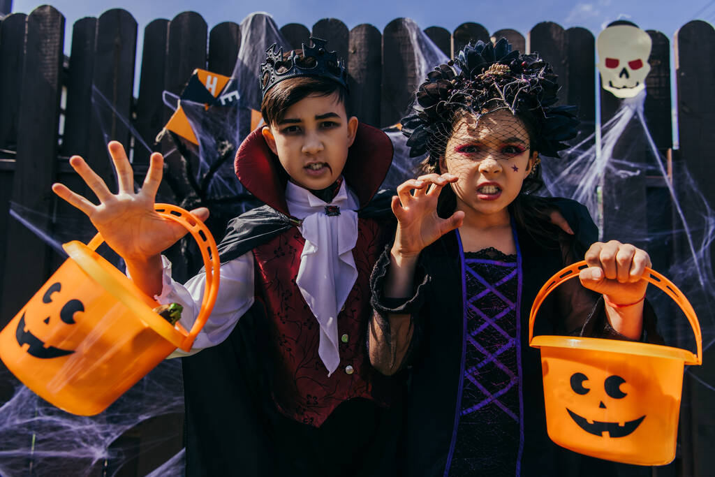 Interrazziale ragazzo e ragazza holding halloween secchi e smorfie a macchina fotografica vicino recinto in cortile  - Foto, immagini