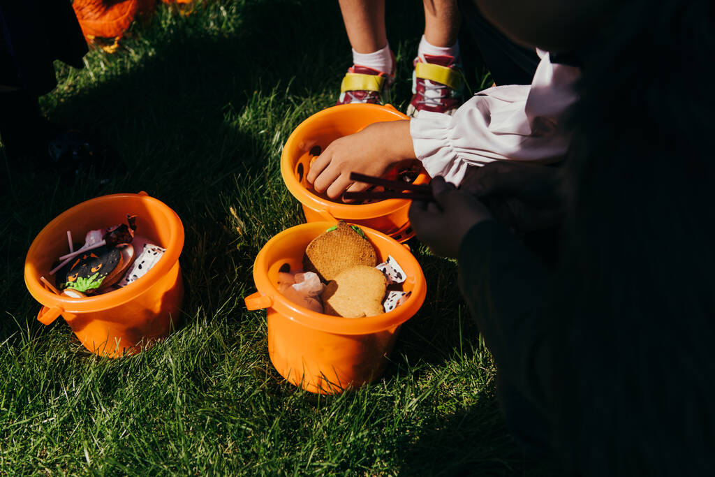 Vista recortada del niño tomando caramelos del cubo durante la fiesta de halloween al aire libre  - Foto, Imagen