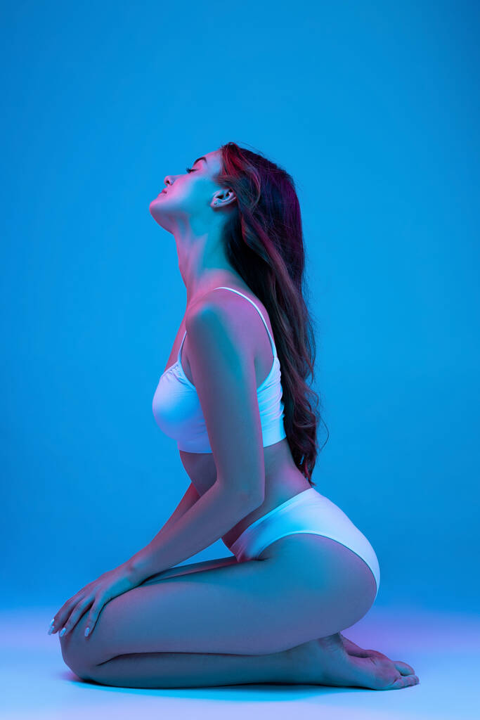 Něžnost. Mladá smyslná žena ve spodním prádle sedí na podlaze izolované nad modrým pozadím v neonovém světle. Wellness, wellness, fitness a estetický kosmetický koncept. Přírodní krása - Fotografie, Obrázek