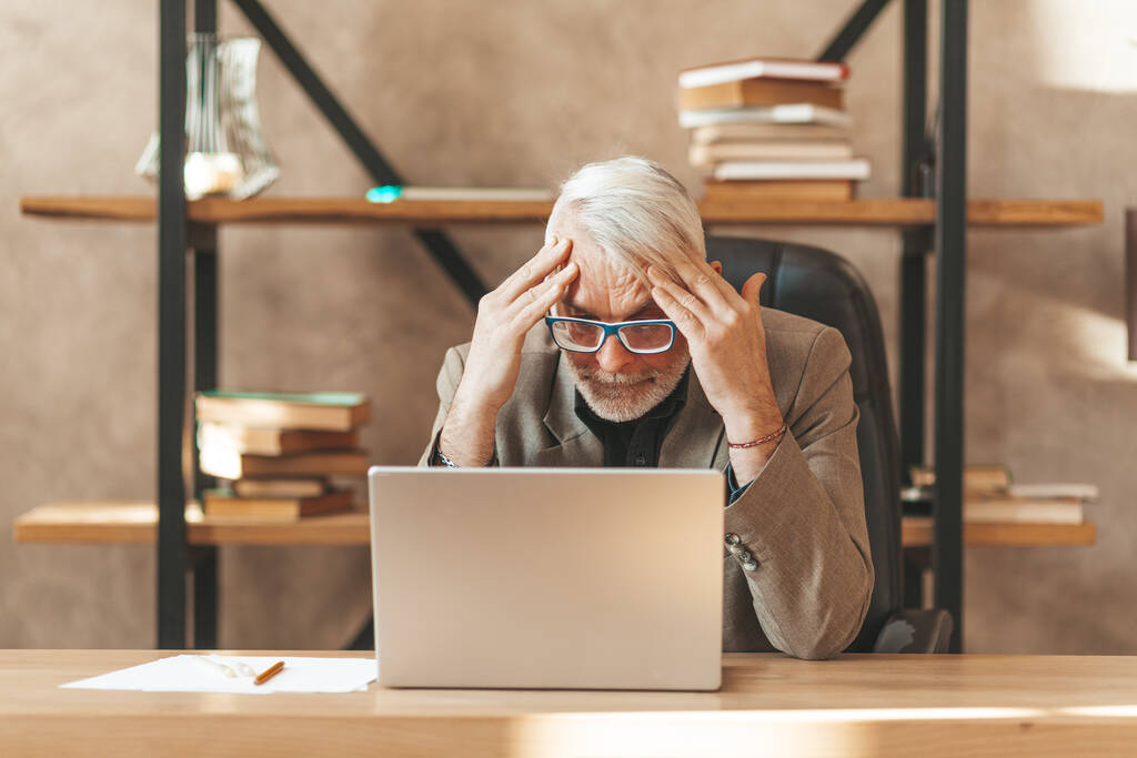 Burnout en la oficina. Un hombre mayor deprimido se sienta en un portátil, sosteniendo su cabeza. Incomprensión y desesperación. - Foto, Imagen