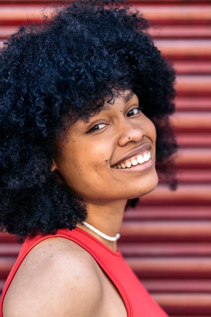 Beautiful young black woman with afro hair smiling and looking at camera against red background. - Photo, Image