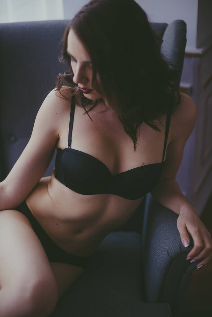 Uma jovem garota atraente com cabelo escuro em lingerie preta com maquiagem à noite sentada em uma poltrona cinza no quarto. beleza e moda - Foto, Imagem