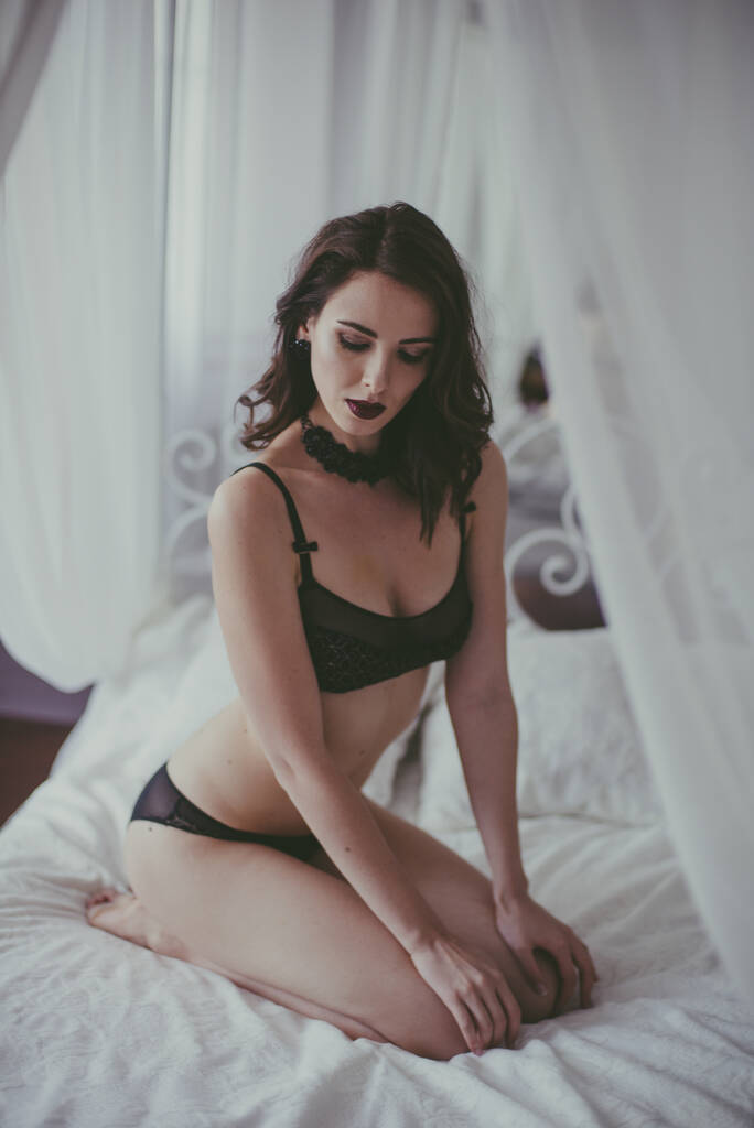 Een jong aantrekkelijk meisje met donker haar in zwarte lingerie met avondmake-up en sieraden en een witte badjas op een wit bed in de slaapkamer. schoonheid en mode - Foto, afbeelding