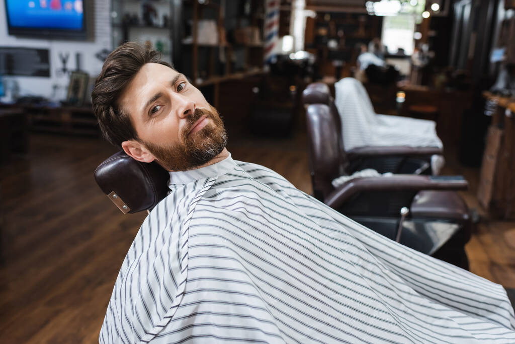 bearded man in hairdressing cape looking at camera in blurred beauty salon - Photo, Image