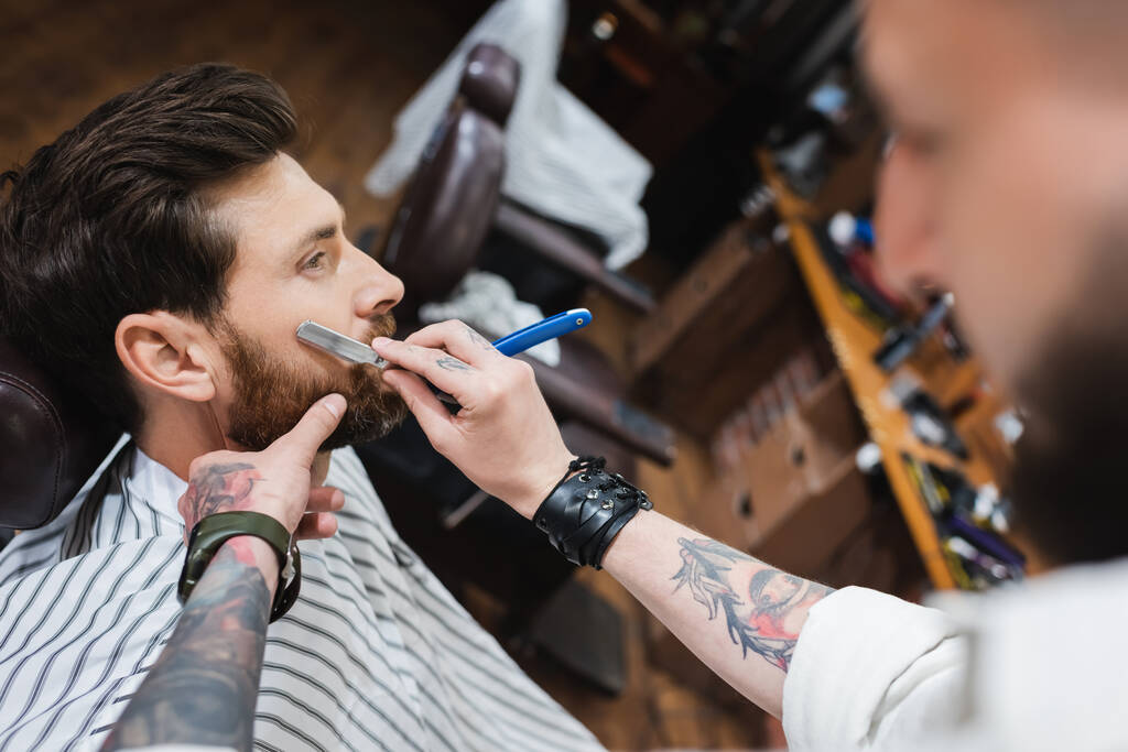 размытый парикмахер в кожаном браслете бритья клиента с прямой бритвой - Фото, изображение