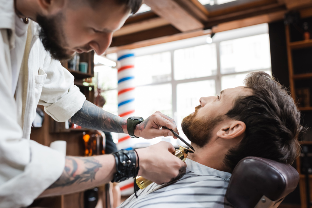 uomo barbuto bruna vicino parrucchiere che lavora con pettine e tagliacapelli - Foto, immagini