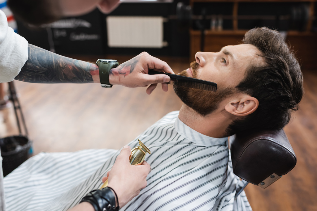 Κουρέας με τατουάζ που κρατά κουρέα μαλλιών και χτένισμα γενειάδα του ανθρώπου στο κουρείο - Φωτογραφία, εικόνα