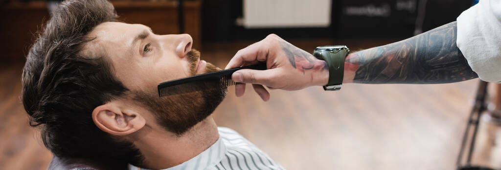 Kol saatinde dövmeli kuaför müşterinin sakalını, afişini tarıyor. - Fotoğraf, Görsel