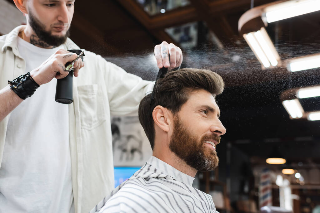 happy brunette man near barber applying hair spray in beauty salon - Photo, Image