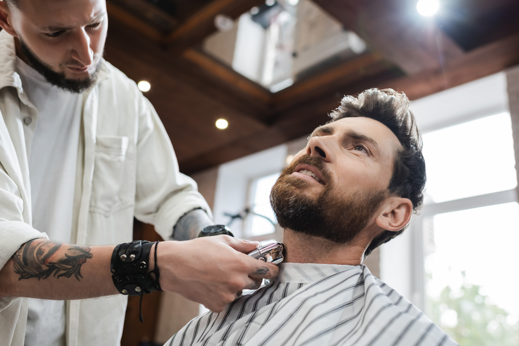 bajo ángulo vista de peluquero afeitado cuello de hombre barbudo con afeitadora eléctrica - Foto, imagen