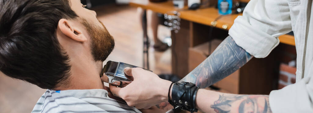 tätowierter Friseur rasiert Hals eines brünetten Mannes mit Rasiermesser, Banner - Foto, Bild