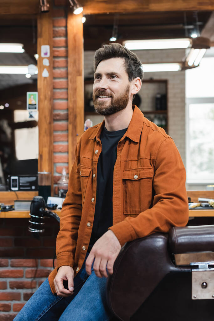 uomo barbuto sorridente seduto sulla poltrona parrucchiere vicino a specchi sfocati in barbiere - Foto, immagini