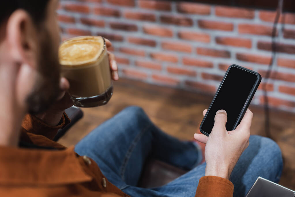 částečný pohled na rozmazaného muže se sklenicí cappuccina držícího smartphone s prázdnou obrazovkou - Fotografie, Obrázek