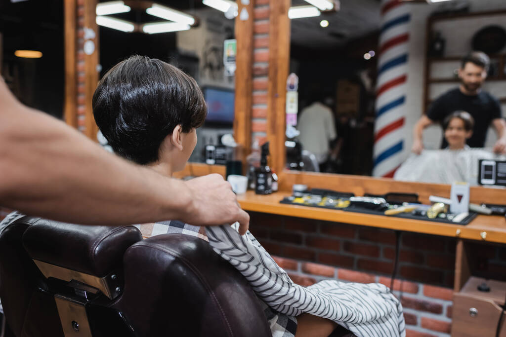 перукар кладе перукарську накидку на підлітка-клієнта, який сидить у кріслі біля розмитих дзеркал
 - Фото, зображення