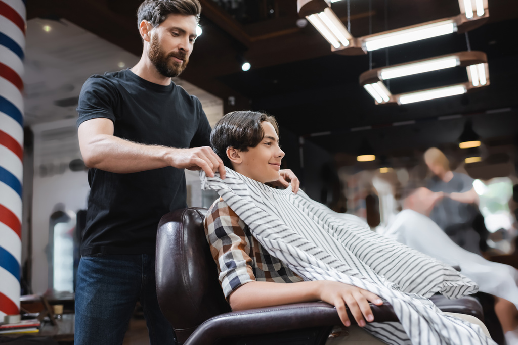 barbero barbudo poniendo capa de peluquería en cliente adolescente sonriendo en sillón - Foto, imagen