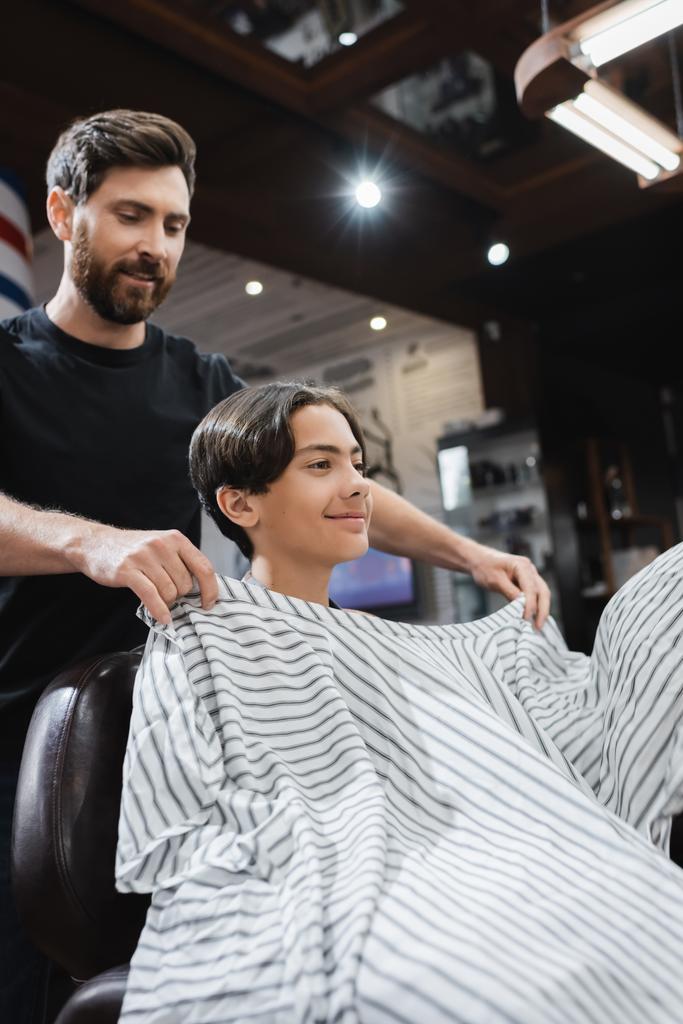 bearded hairstylist putting hairdressing cape on smiling teenager in barbershop - Photo, Image