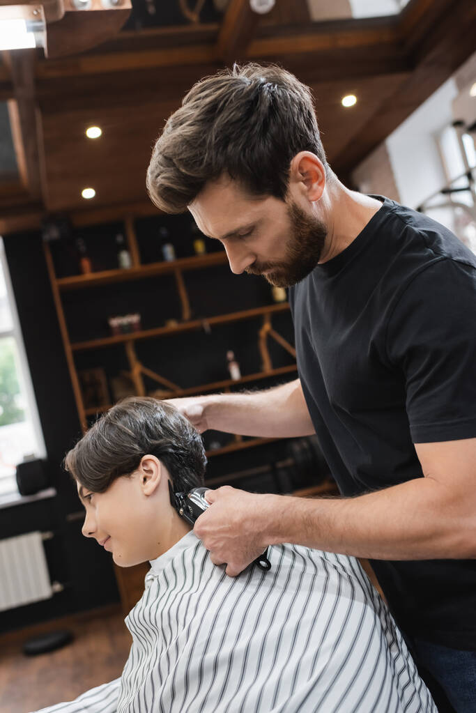 πλαϊνή άποψη του γενειοφόρου κουρέα κούρεμα μαλλιά του έφηβος αγόρι σε hairdressing ακρωτήριο - Φωτογραφία, εικόνα
