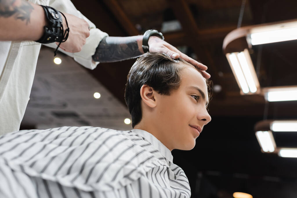 Niedrigwinkel-Ansicht tätowierter Friseur berührt Haare eines brünetten Typen im Friseursalon - Foto, Bild