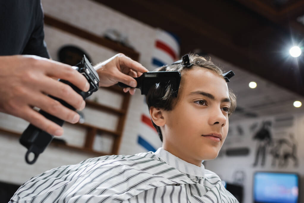 dospívající chlapec s sponkami do vlasů v blízkosti holiče česání jeho vlasy a držení střihače vlasů - Fotografie, Obrázek