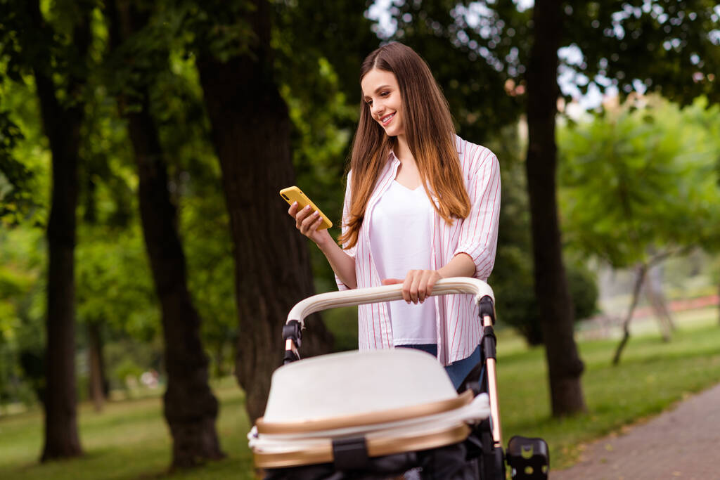 Photo of positive cute mother dressed casual clothes walking buggy little son typing modern device outdoors backyard. - Photo, Image