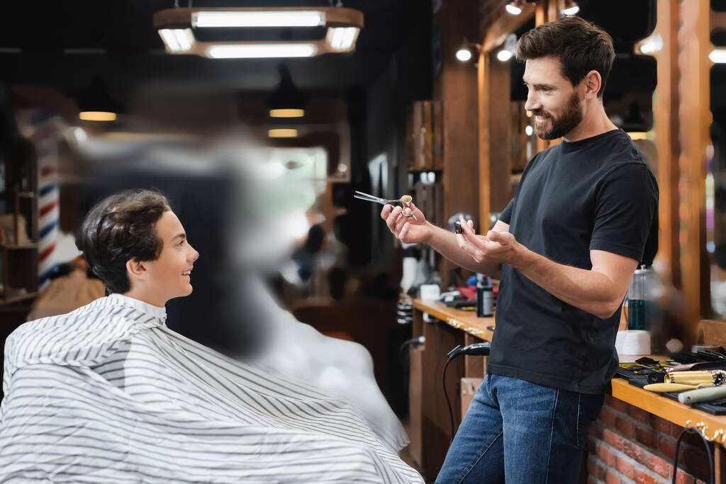 Pozytywny nastolatek chłopiec w pelerynie patrząc na fryzjera z nożyczkami i grzebień w salonie piękności  - Zdjęcie, obraz