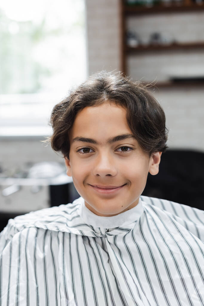 Усміхнений підліток в перукарській накидці дивиться на камеру в салоні краси
  - Фото, зображення