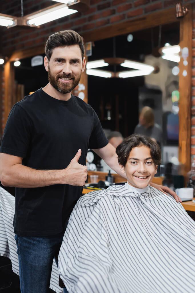 Pozytywny fryzjer pokazując jak gest i patrząc na kamerę w pobliżu nastolatka w pelerynie fryzjerskiej  - Zdjęcie, obraz