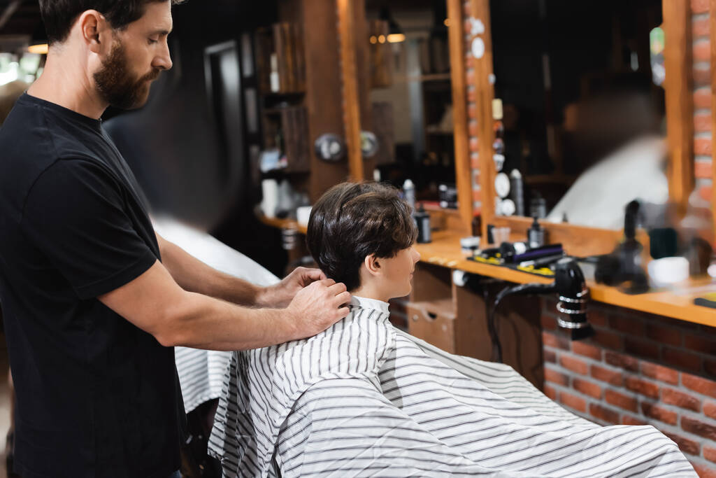 Вид на парикмахера в плаще на мальчика-подростка в салоне красоты  - Фото, изображение