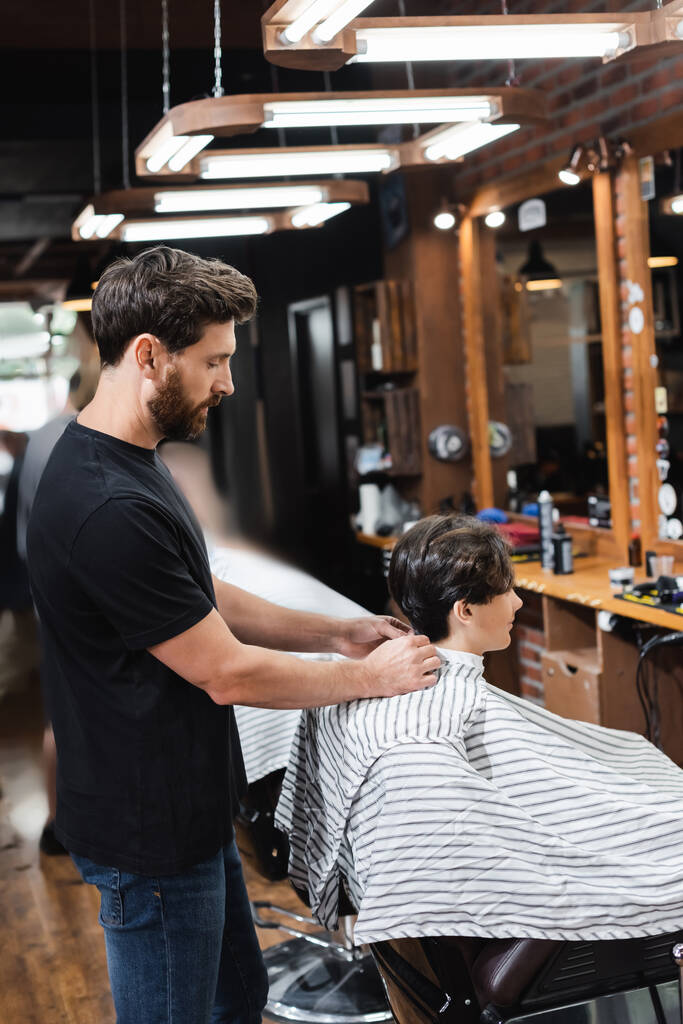 Side view of barber wearing hairdressing cape on teen boy in barbershop  - Photo, Image