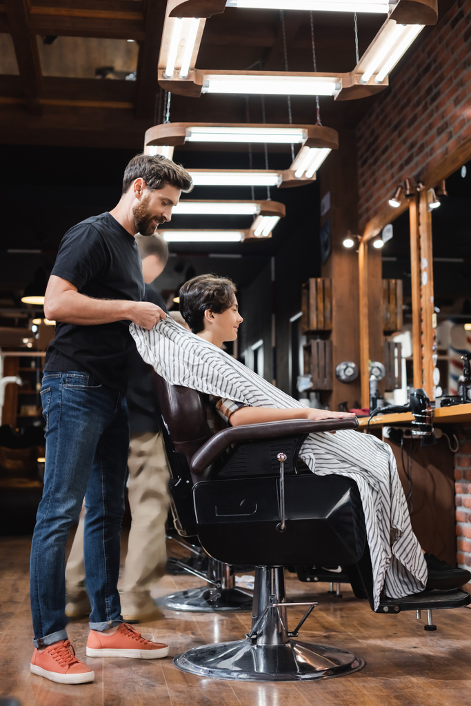 Vista lateral do cabeleireiro decolando capa de cliente adolescente no salão de beleza  - Foto, Imagem