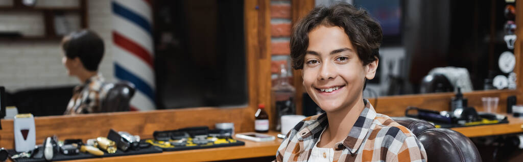 Adolescente sonriendo a la cámara mientras está sentado en el sillón en la barbería, pancarta  - Foto, imagen