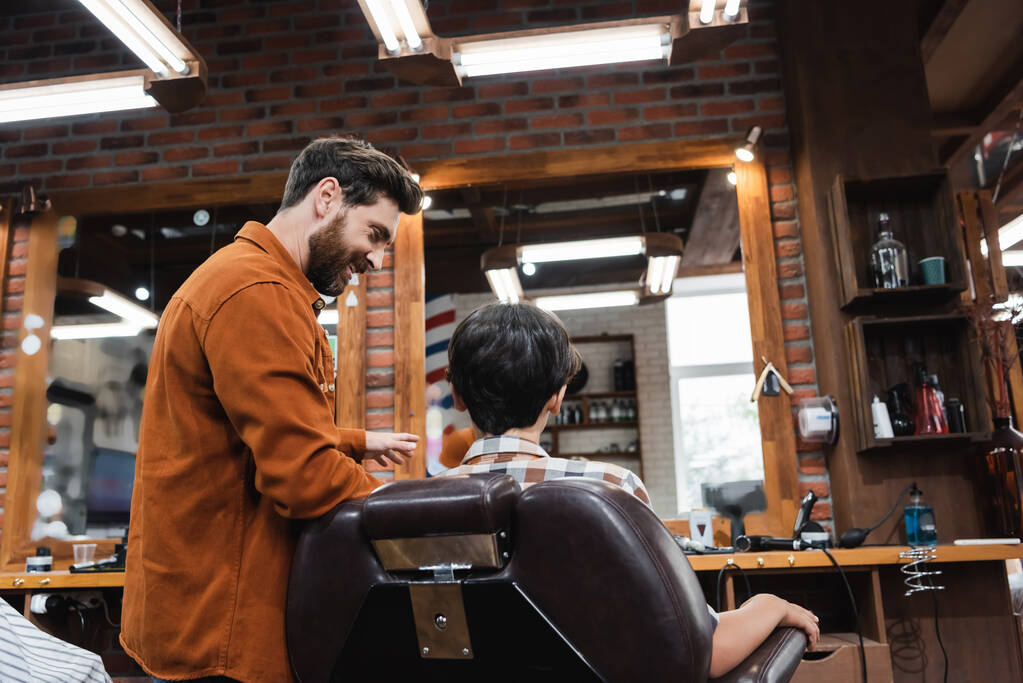 Sorridente barbiere parlare con adolescente seduto in poltrona in barbiere  - Foto, immagini