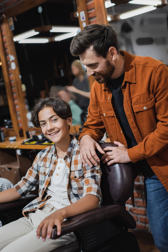 Веселий підліток клієнт дивиться на камеру, сидячи в кріслі біля перукарні в перукарні
  - Фото, зображення
