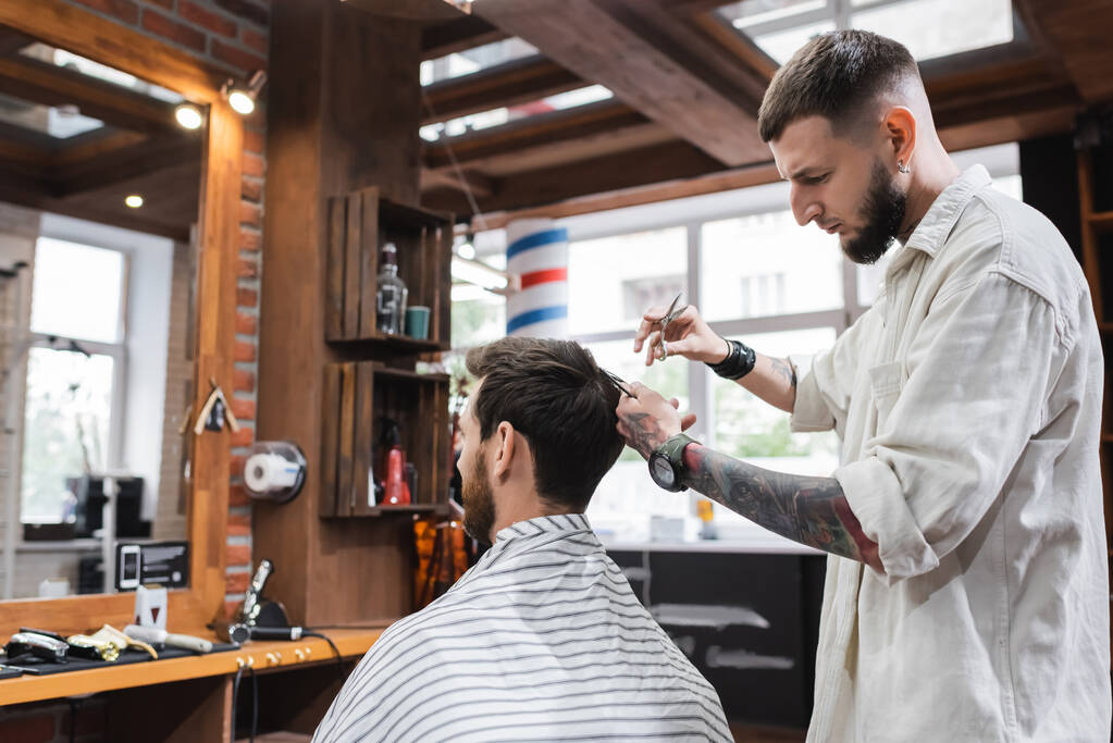 Tätowierter Friseur kämmt Haare seiner Kundin und hält Schere im Friseurladen  - Foto, Bild