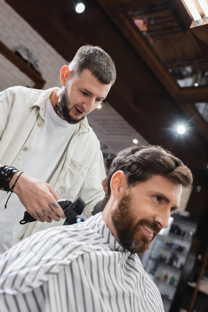 Coiffeur tenant une tondeuse à cheveux et parlant à un client souriant dans un salon de beauté  - Photo, image