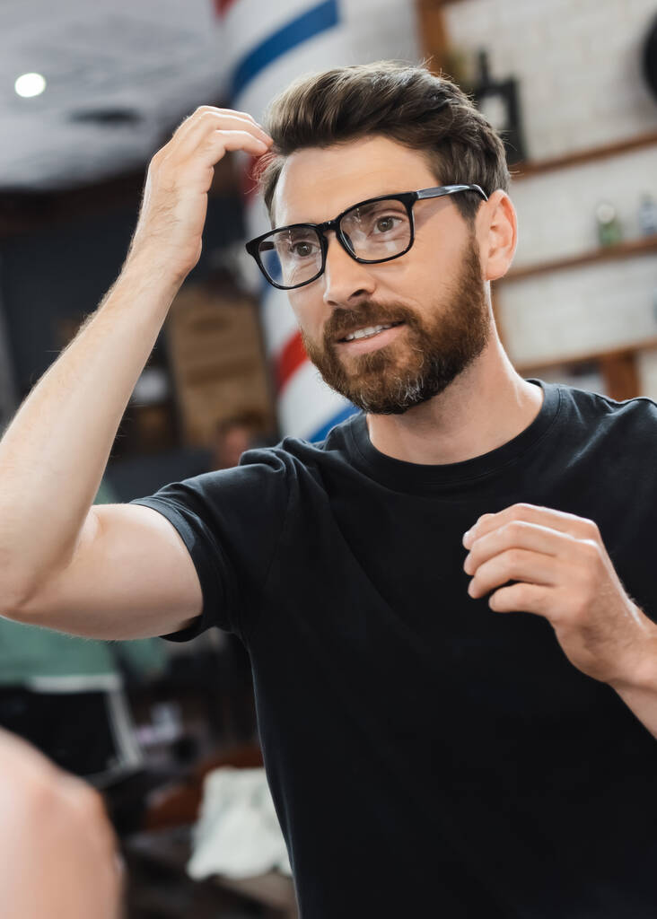Усміхнений чоловік в окулярах торкається волосся біля дзеркала в перукарні
  - Фото, зображення