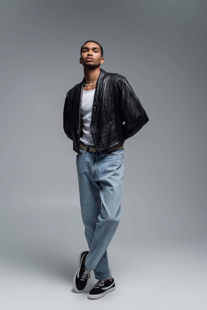 piena lunghezza di elegante uomo afroamericano in abito autunnale in posa con gambe incrociate sul grigio  - Foto, immagini