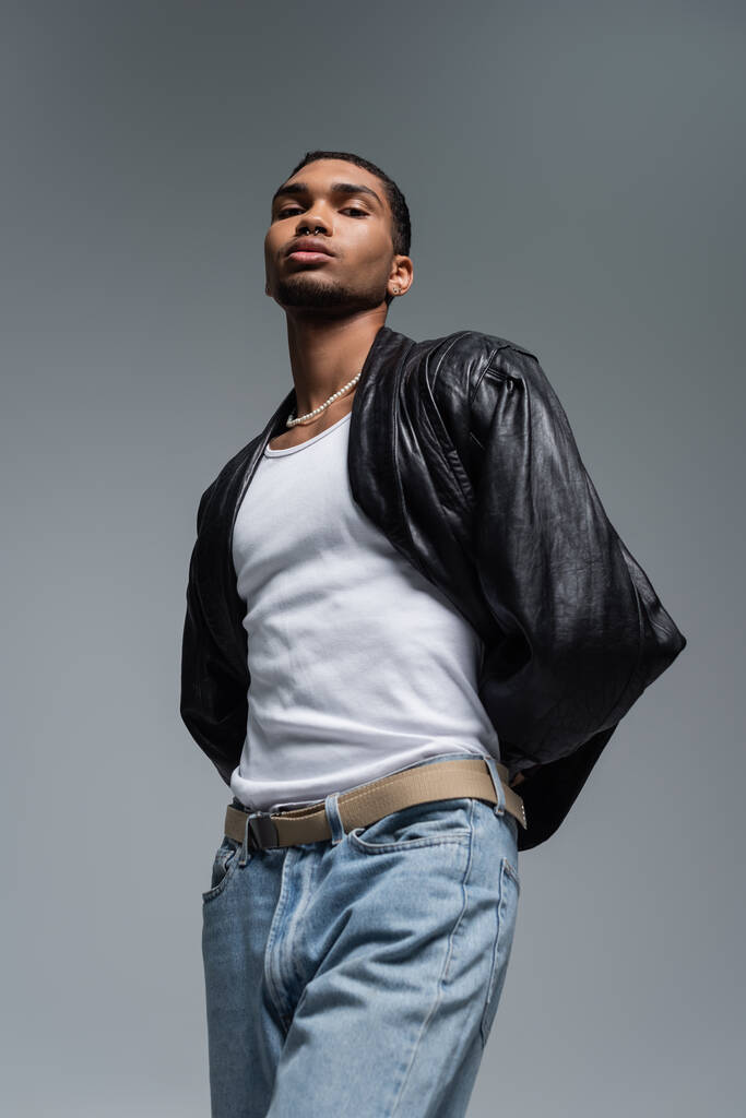 nízký úhel pohledu na mladého afrického Američana muže v džínách a kožené bundě pózující izolované na šedé  - Fotografie, Obrázek