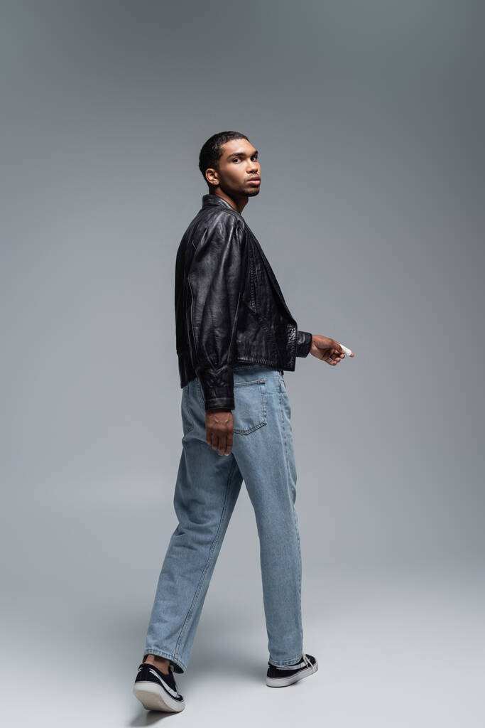 full length of young αφροαμερικανός άνδρας με δερμάτινο μπουφάν περπάτημα σε γκρι  - Φωτογραφία, εικόνα