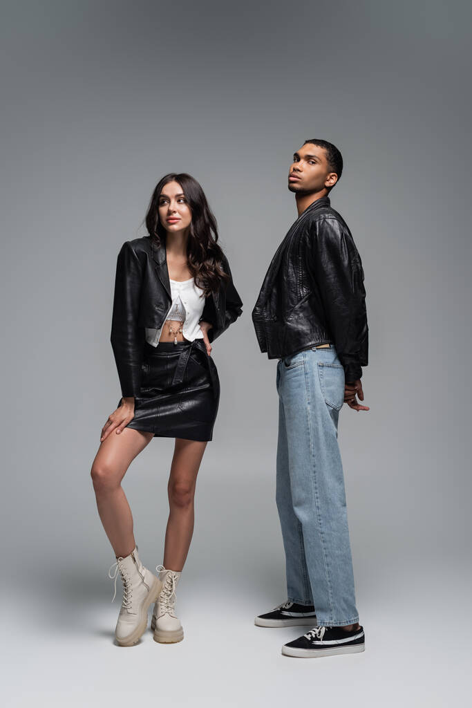 full length of stylish woman in leather outfit and boots posing with young african american man on grey - Photo, Image
