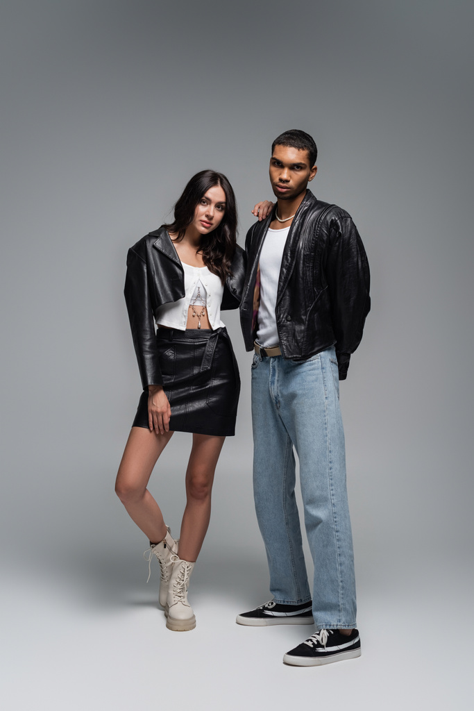 full length of young woman in leather outfit and boots posing with stylish african american man on grey - Photo, Image