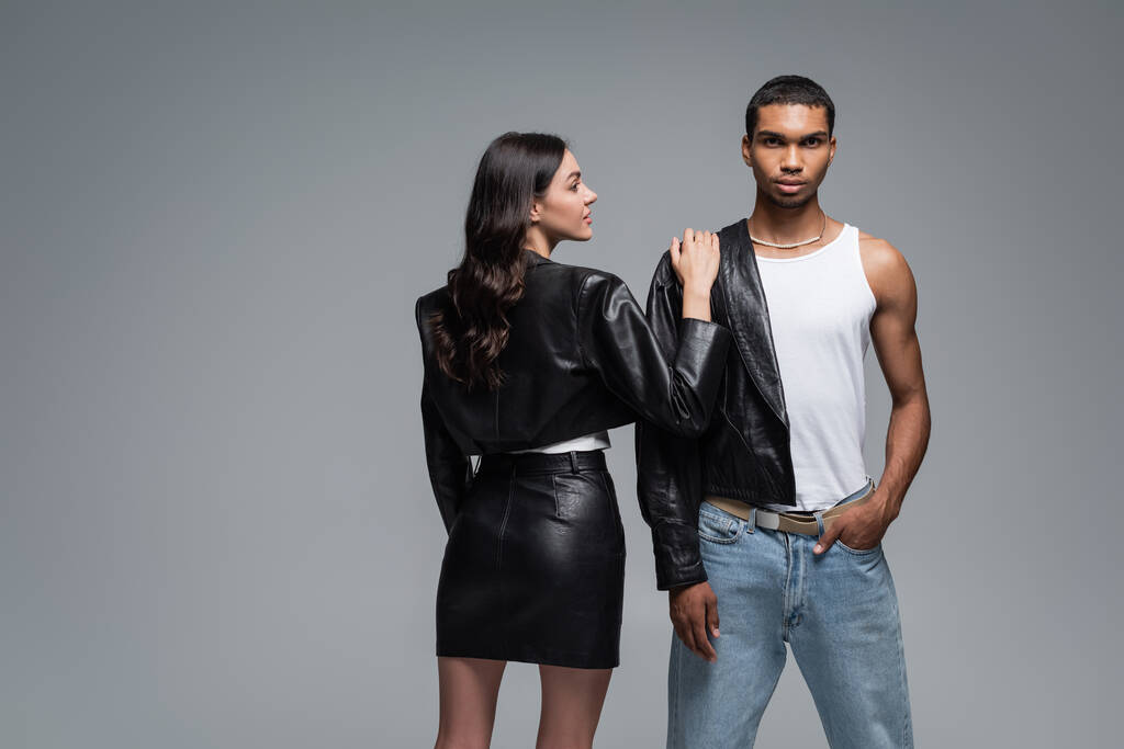 young woman in leather outfit hugging stylish african american man posing with hand in pocket isolated on grey - Photo, Image
