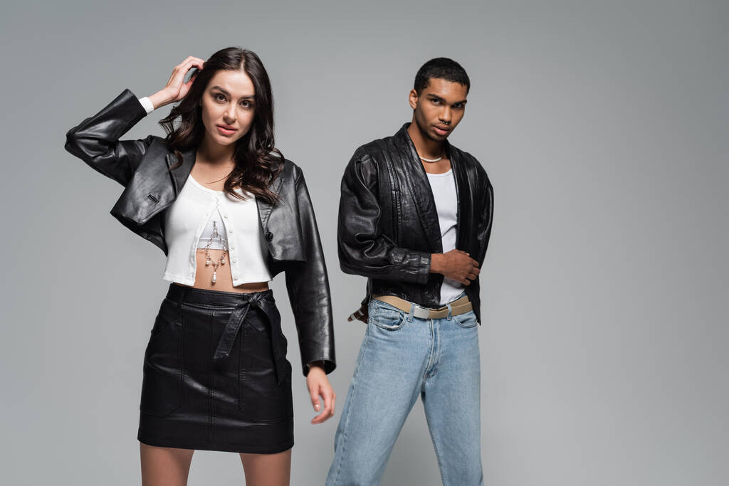 young and stylish multiethnic couple in leather jackets posing isolated on grey - Photo, Image