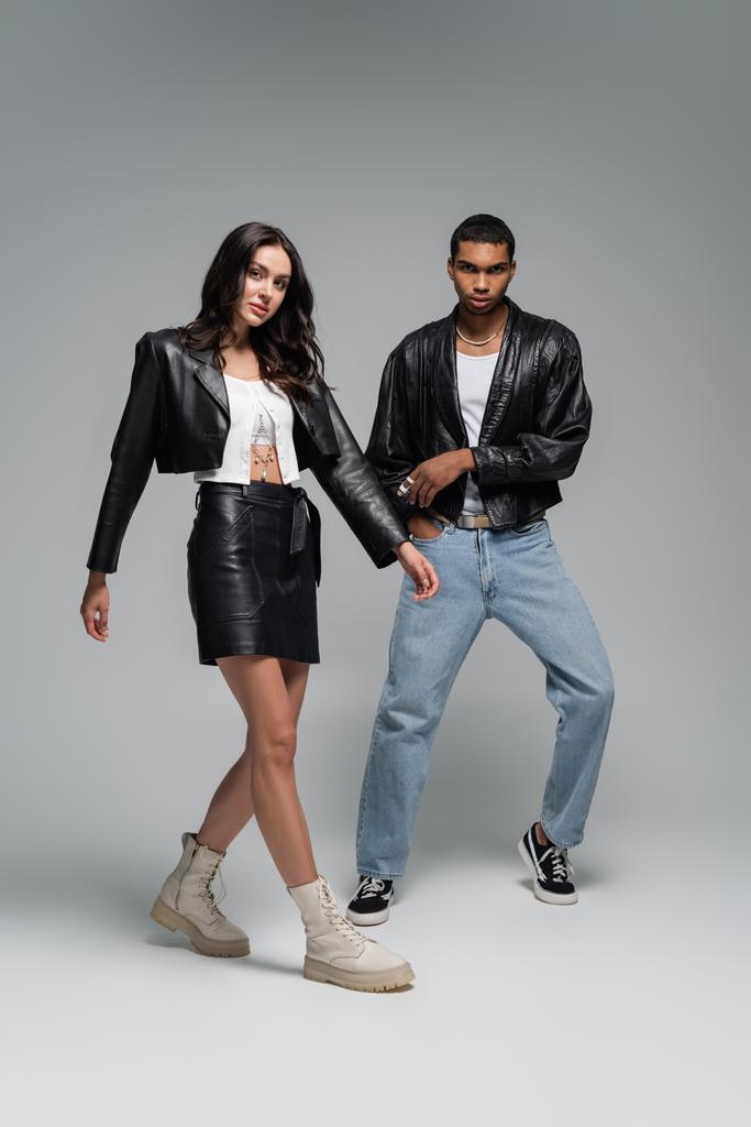 full length of young and stylish multiethnic couple in leather jackets posing on grey - Photo, Image