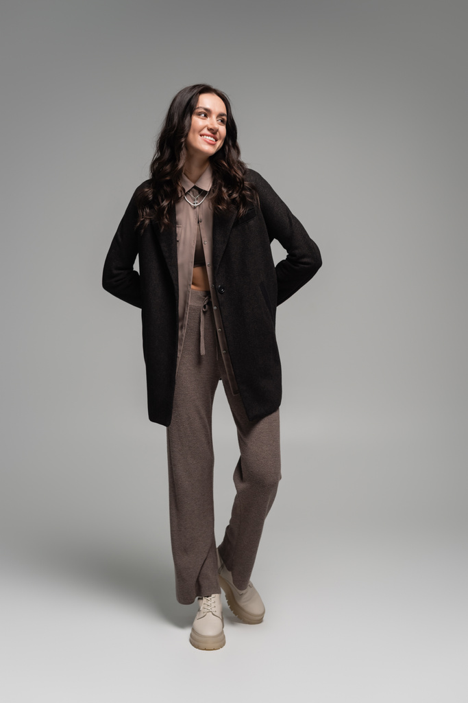 volledige lengte van gelukkige jonge vrouw in zwarte blazer en gebreide broek poseren op grijs - Foto, afbeelding
