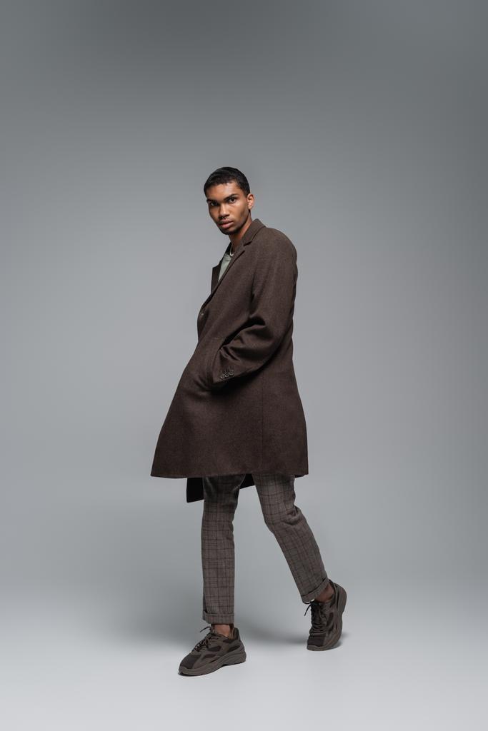 teljes hossza fiatal afro-amerikai férfi őszi gyapjú kabát néz kamera és pózol kéz a zsebben szürke  - Fotó, kép
